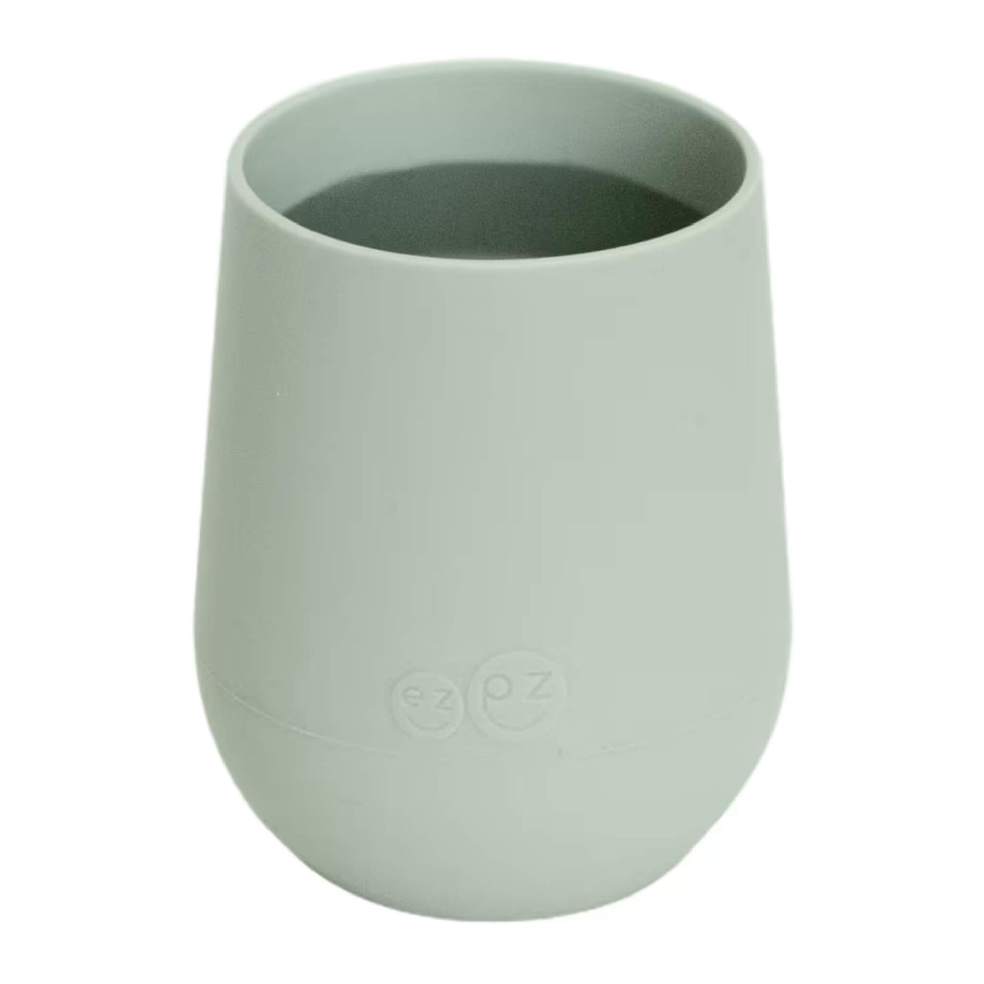 A sage green silicone training cup shown filled with milk, designed for toddlers learning to drink from an open cup.