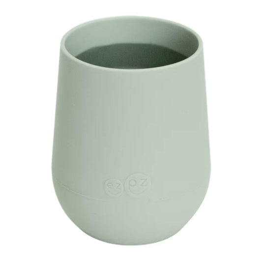 A sage green silicone training cup shown filled with milk, designed for toddlers learning to drink from an open cup.