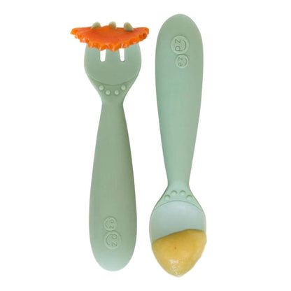 Sage green toddler training fork holding a slice of carrot beside a matching spoon filled with puree.