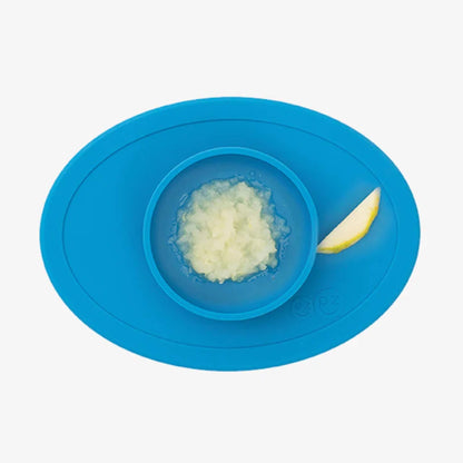 Blue silicone placemat with a small bowl containing mashed food and a slice of pear resting on the mat beside it.