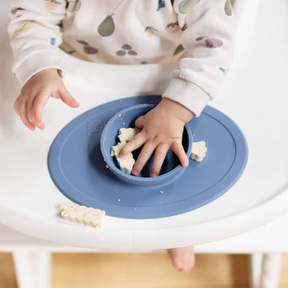 Baby sitting in a highchair pressing food into a silicone suction bowl on a placemat in indigo colour.