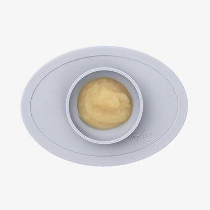 A grey silicone placemat with a small round bowl filled with smooth yellow puree, shown from above.