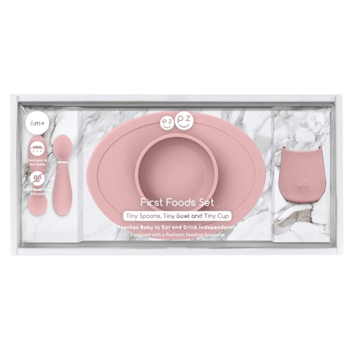 A baby first foods set in a clear plastic tray with a white marble-patterned insert. The set includes a dusty pink silicone bowl with a matching rim, two small pink spoons, and a small pink cup.