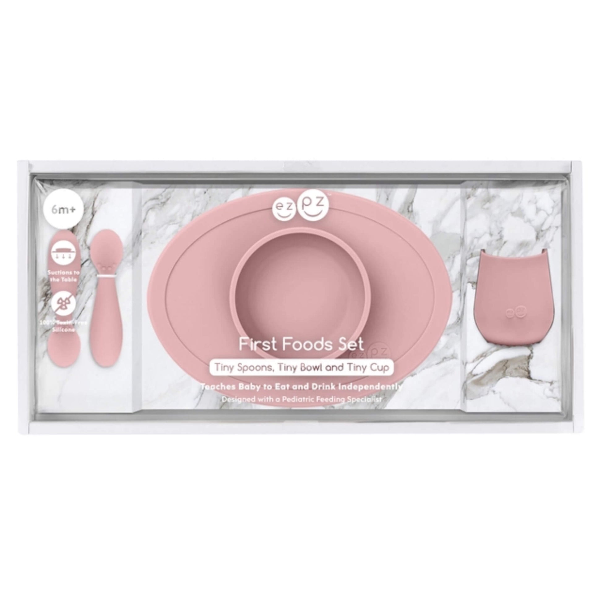A baby first foods set in a clear plastic tray with a white marble-patterned insert. The set includes a dusty pink silicone bowl with a matching rim, two small pink spoons, and a small pink cup.
