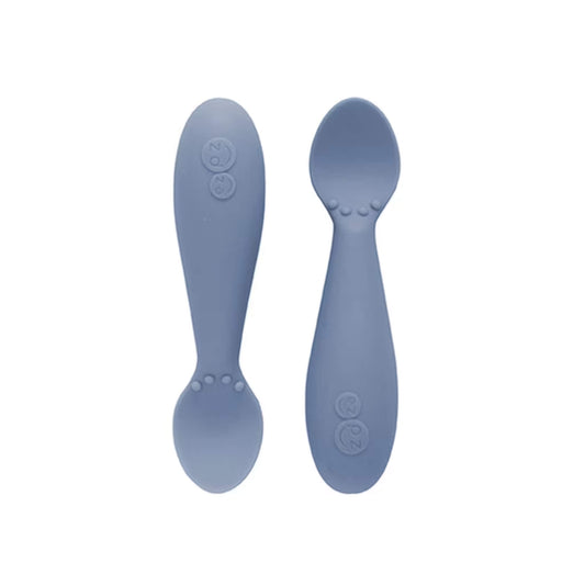 Ezpz Tiny Spoons in indigo blue, a pair of silicone infant training spoons with shallow heads and textured bumps near the base.