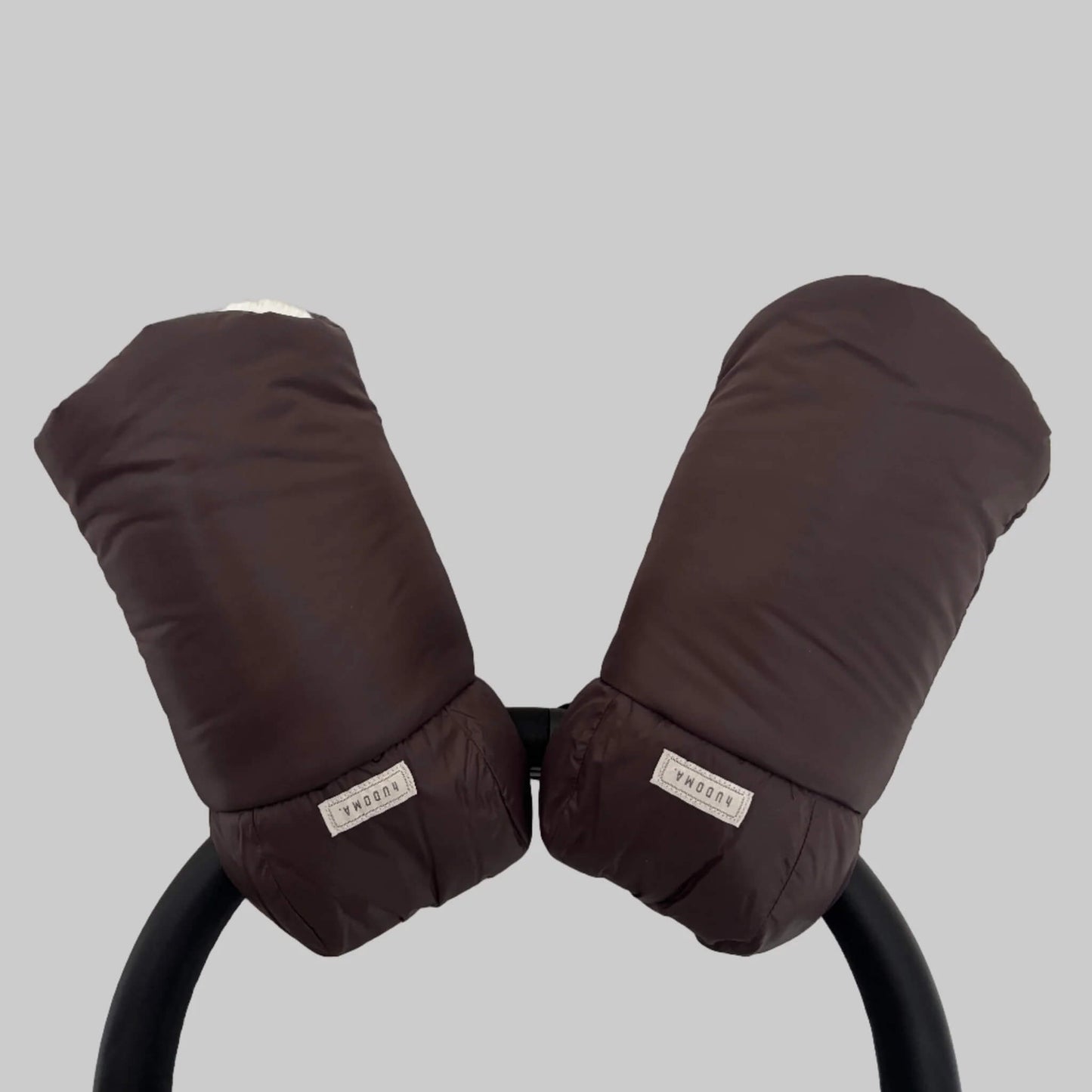 Brown hUDOMA Broadway Pram Gloves attached to a stroller handle, showing both gloves together with visible brand labels and soft faux fur lining inside.
