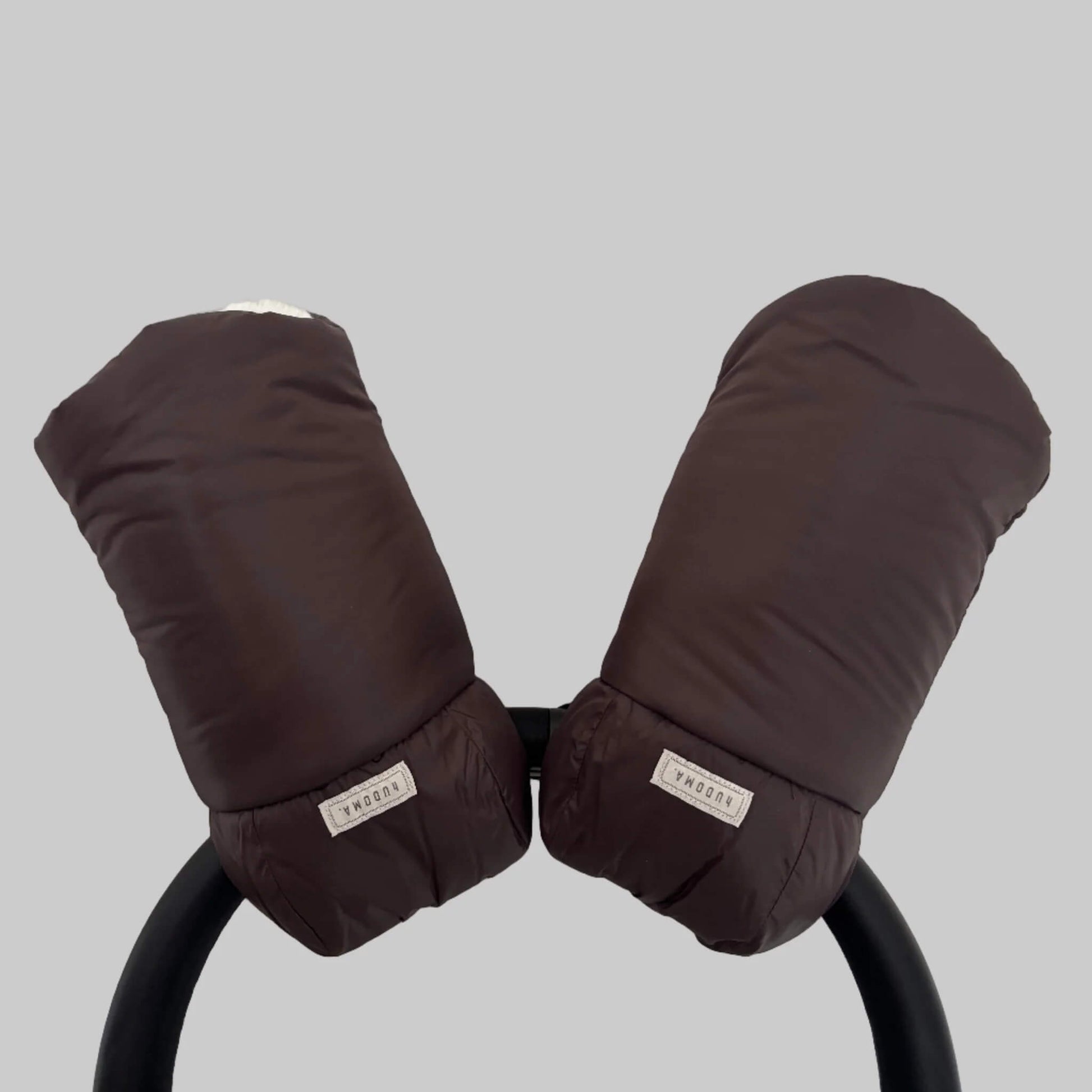 Brown hUDOMA Broadway Pram Gloves attached to a stroller handle, showing both gloves together with visible brand labels and soft faux fur lining inside.