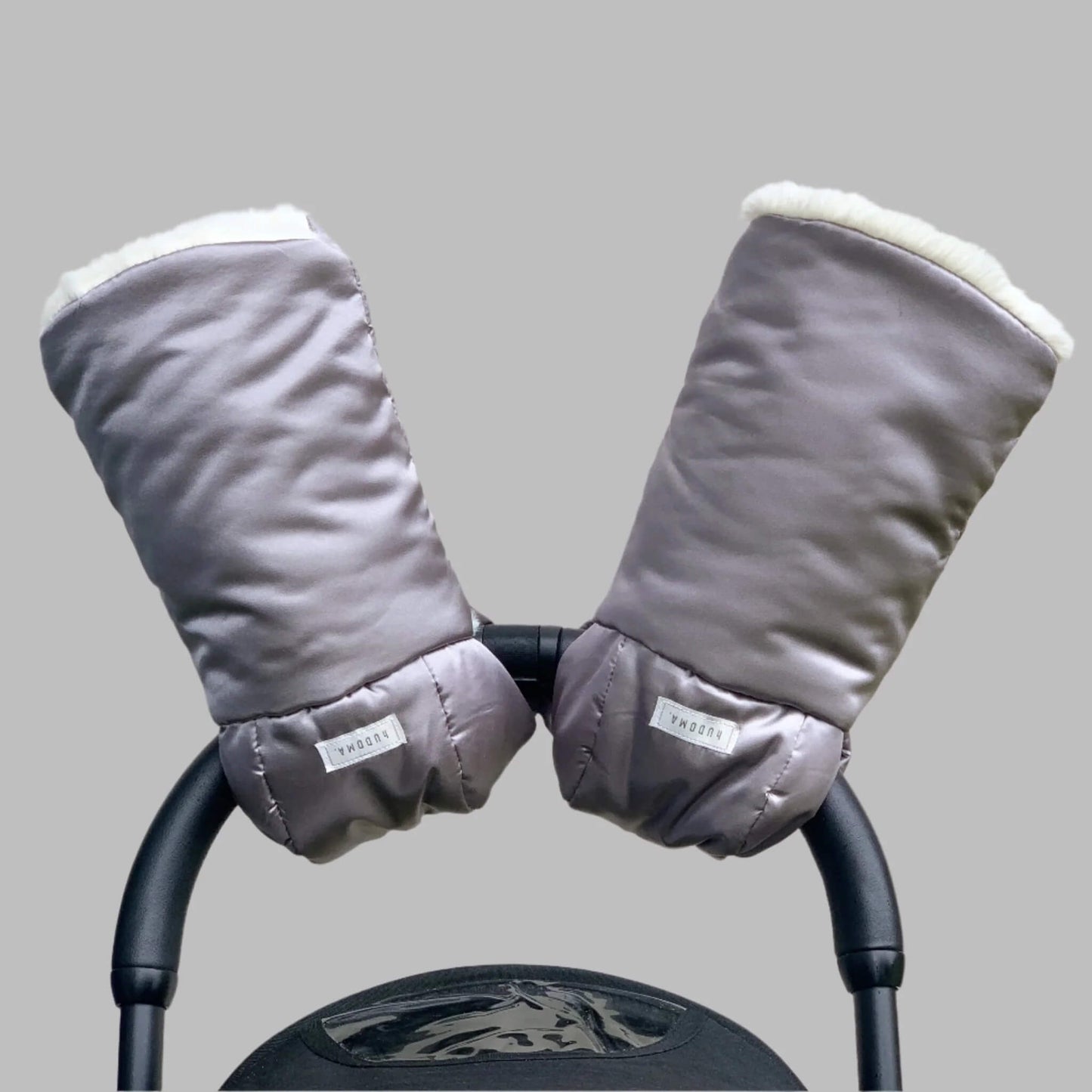 A pair of hUDOMA Pram Gloves in SoHo Silver attached to a black pushchair handle, lined with soft white fleece for comfort and warmth.