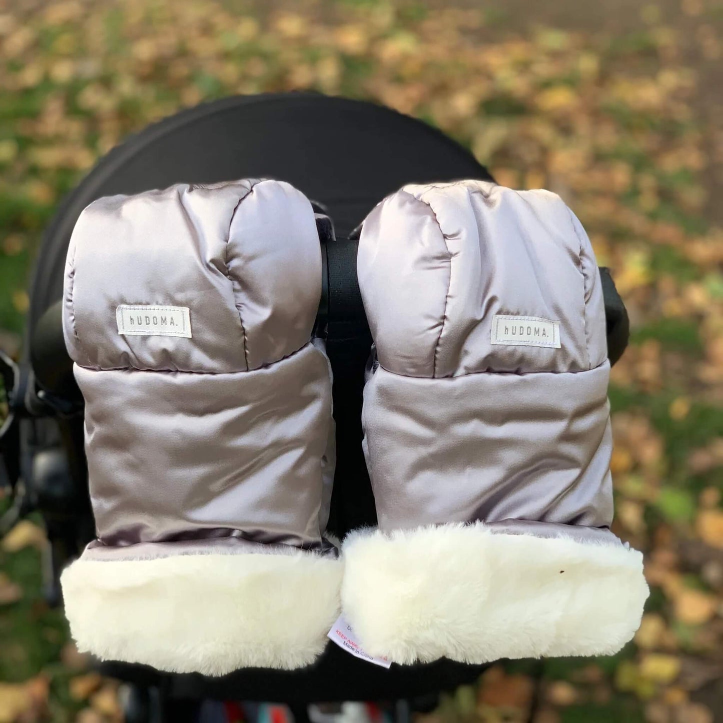 Pram gloves with faux-fur cuffs attached to a stroller handle in an autumn park.