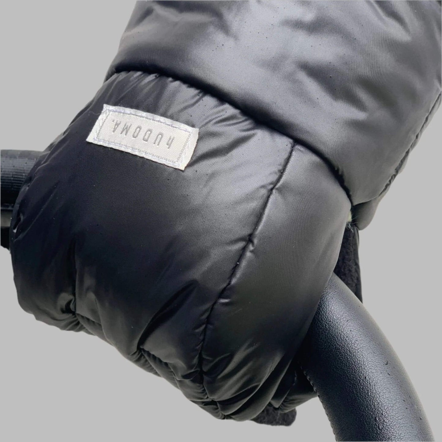 Close-up of a Pram Glove, highlighting its durable black exterior, soft texture, and branded label, securely wrapped around a stroller handle for cold-weather comfort and convenience.